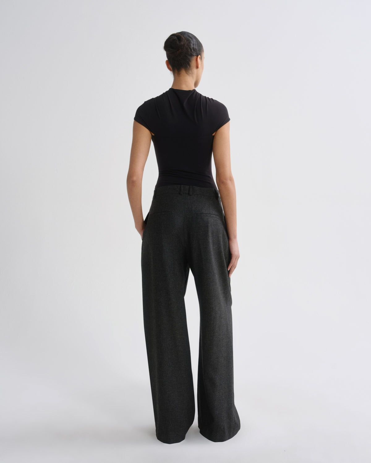 A model viewed from the back wearing a black short-sleeve top and dark charcoal wide-leg trousers, standing against a light gray studio background.
