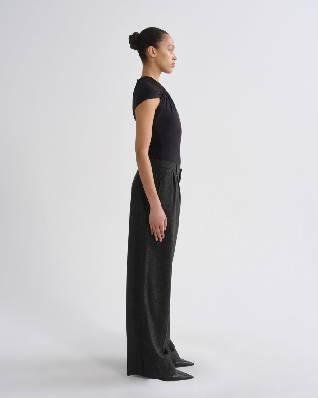 A model in profile wearing a black short-sleeve top and matching black wide-leg trousers, standing against a clean light gray studio background.
