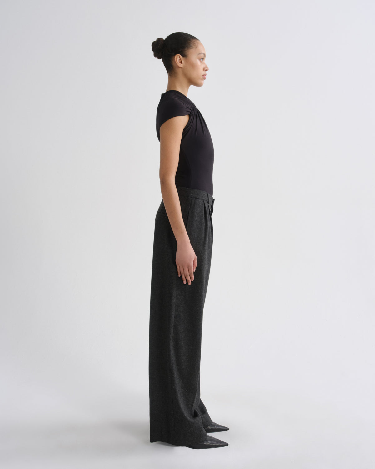 A model in profile wearing a black short-sleeve top and matching black wide-leg trousers, standing against a clean light gray studio background.
