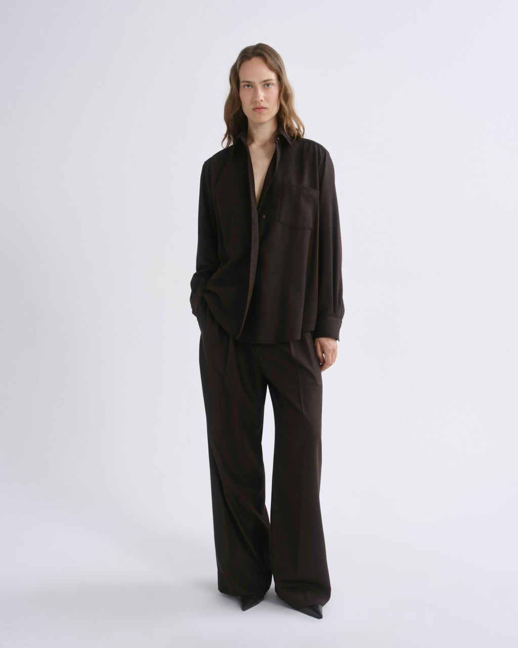A model stands against a grey background wearing a dark brown long-sleeve button-front jacket with a relaxed, oversized fit and matching wide-leg trousers.