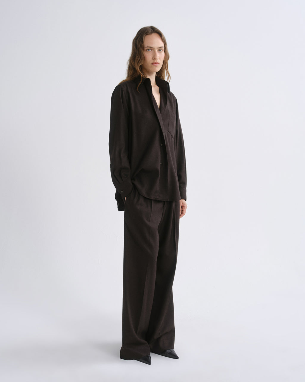 A model stands in a studio wearing a dark brown long-sleeve button-up shirt and matching wide-leg trousers against a grey background.