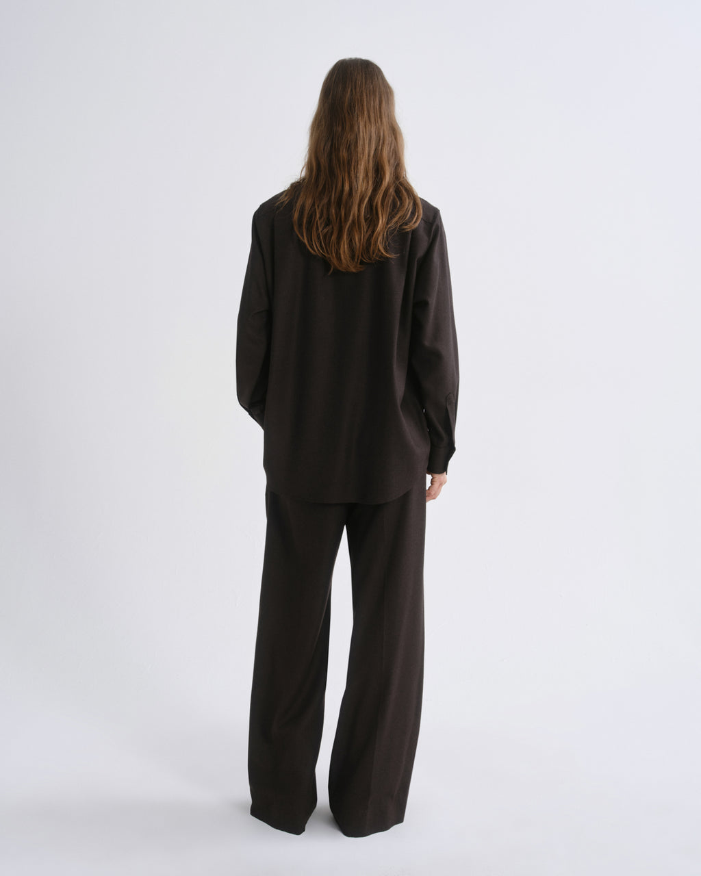 A model viewed from the back wearing a dark brown oversized long-sleeve top and matching wide-leg trousers against a grey background.