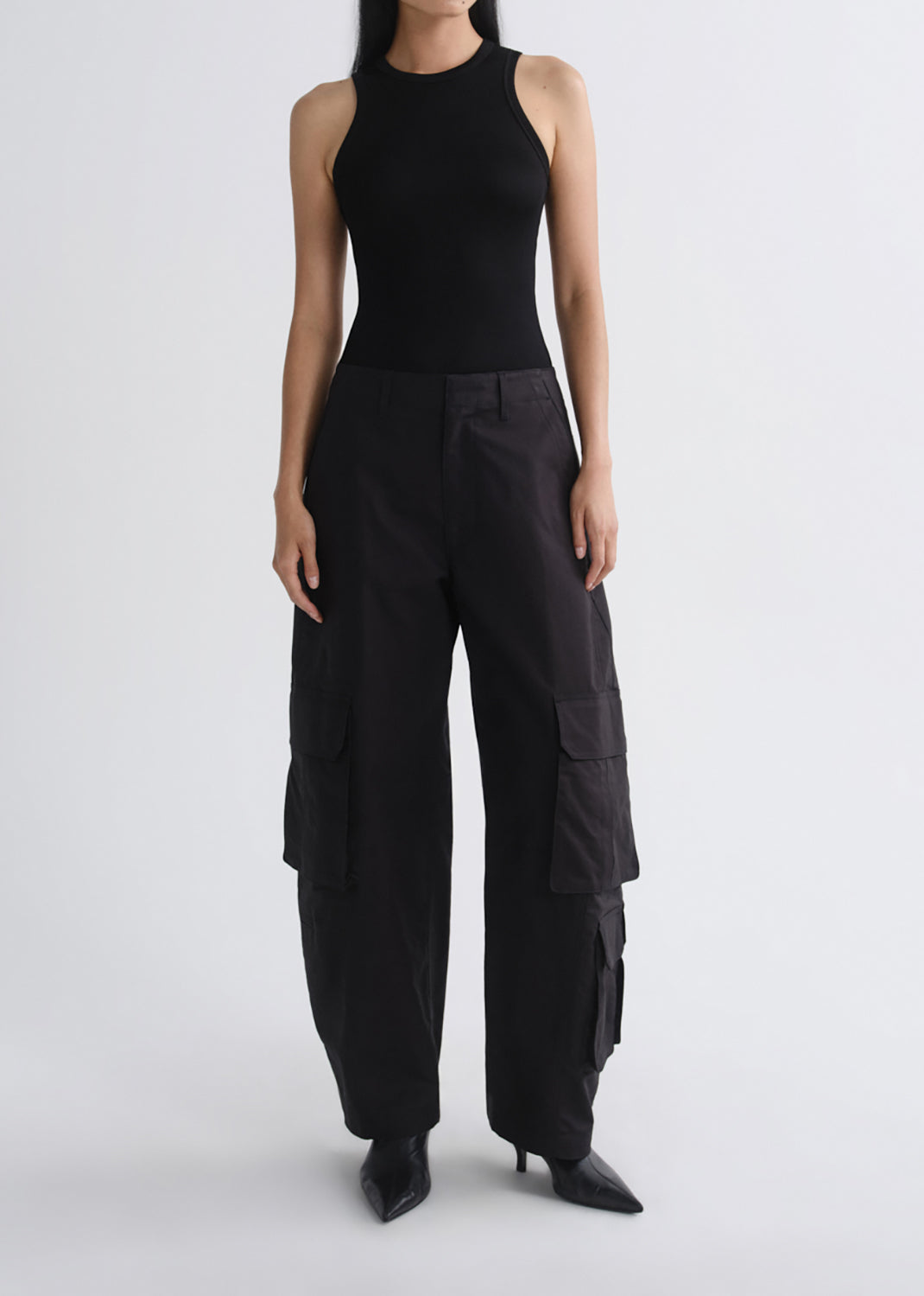 A model wearing a black sleeveless ribbed bodysuit and black wide-leg cargo pants with side flap pockets, paired with black pointed-toe heels, against a grey background.