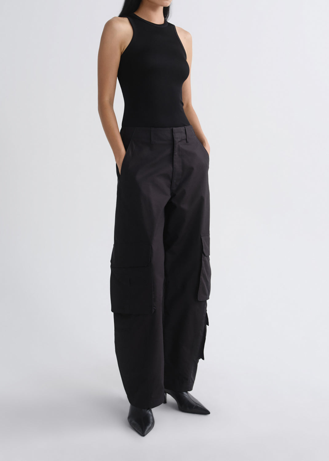 A model wearing a sleeveless black bodysuit and wide-leg black cargo pants with black pointed-toe heels, standing against a grey background.