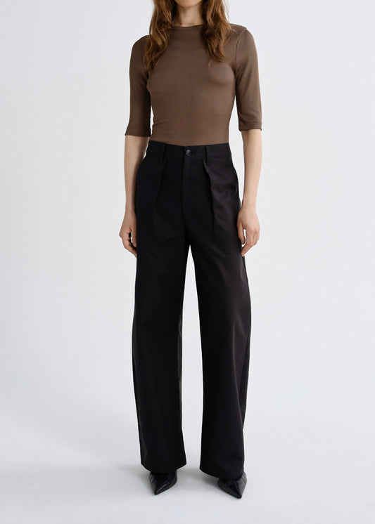A model wearing a brown ribbed three-quarter sleeve top and black wide-leg trousers, standing against a grey background.