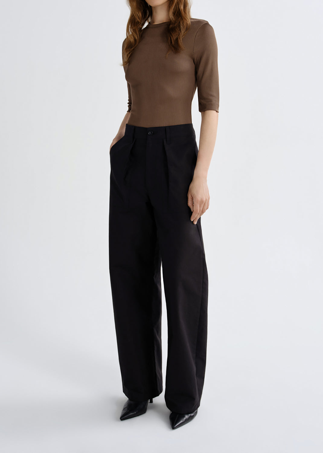 A model wearing a brown short-sleeve mock-neck top and black wide-leg trousers, standing against a grey background.