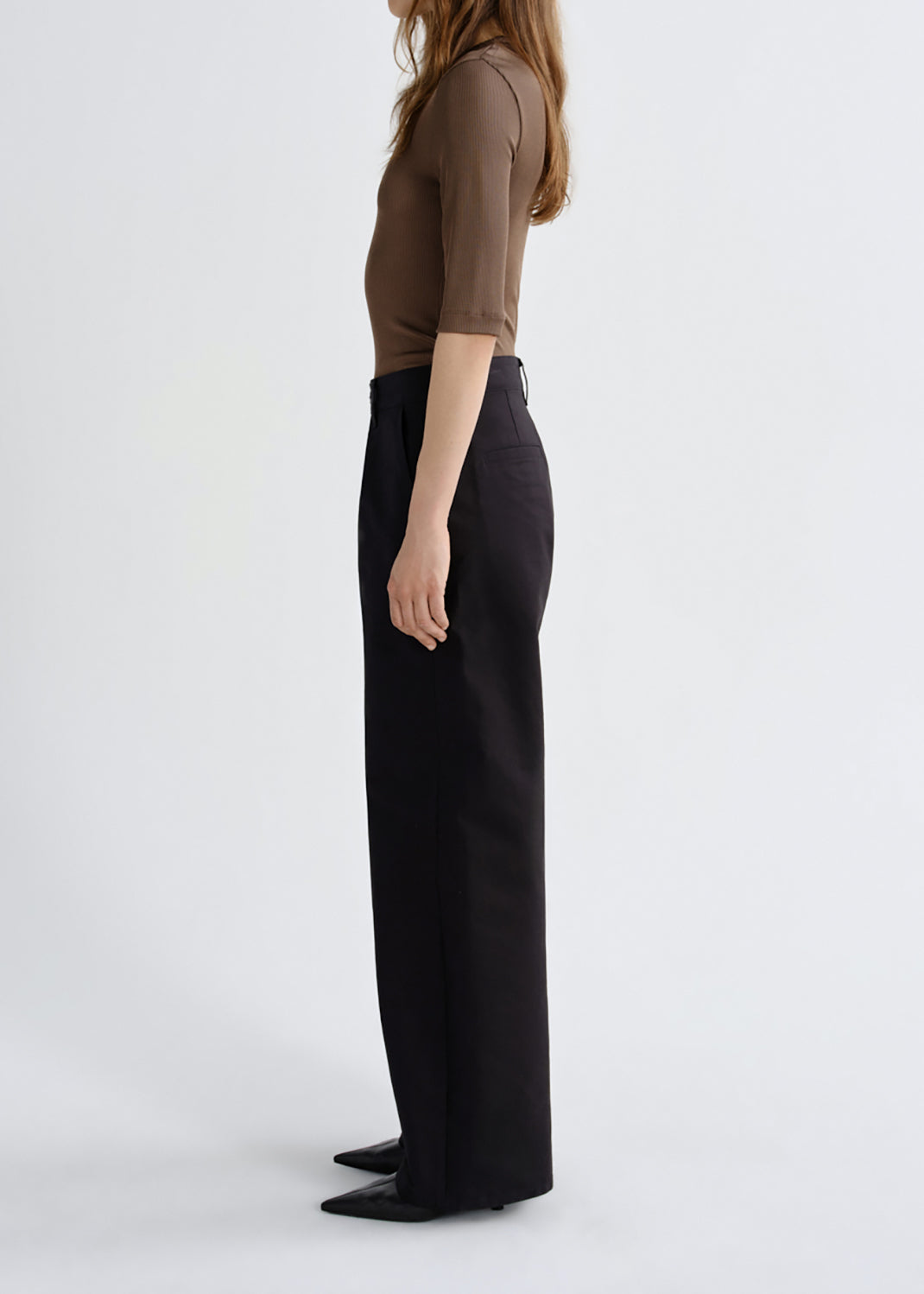 A model shown from the side profile wearing a brown short-sleeve top and black wide-leg trousers against a grey background.
