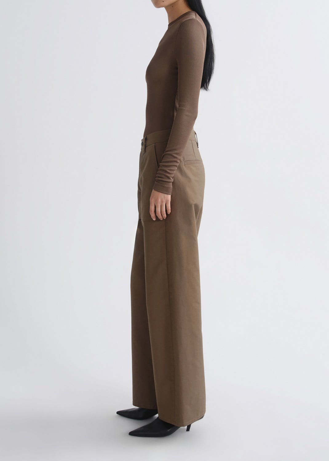 A model shown in profile wearing a brown long-sleeve mock-neck top and matching brown wide-leg trousers, paired with black pointed-toe heels, against a grey background.