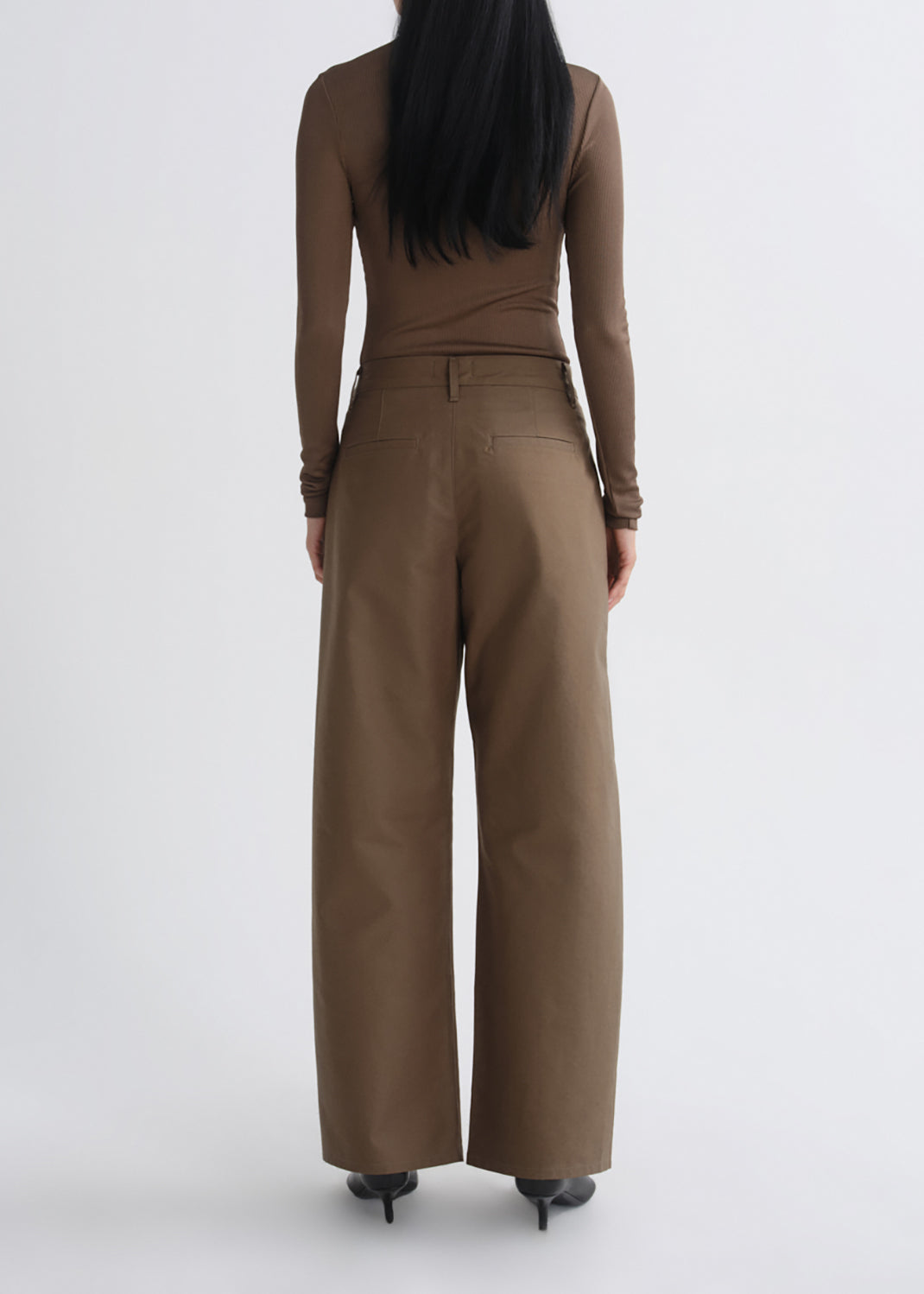 A model viewed from the back wearing a brown long-sleeve top and matching wide-leg trousers, paired with black pointed-toe heels, against a grey background.