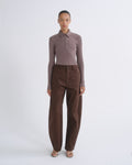 A model stands against a light gray studio background wearing a fitted long-sleeve polo shirt in muted taupe-brown and wide-leg brown trousers.