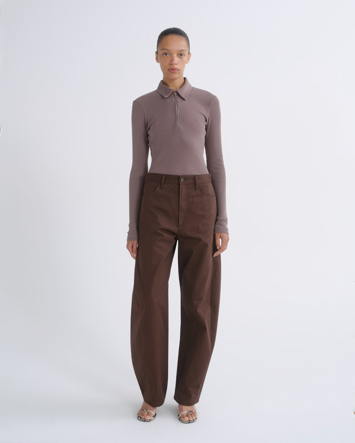 A model stands against a light gray studio background wearing a fitted long-sleeve polo shirt in muted taupe-brown and wide-leg brown trousers.