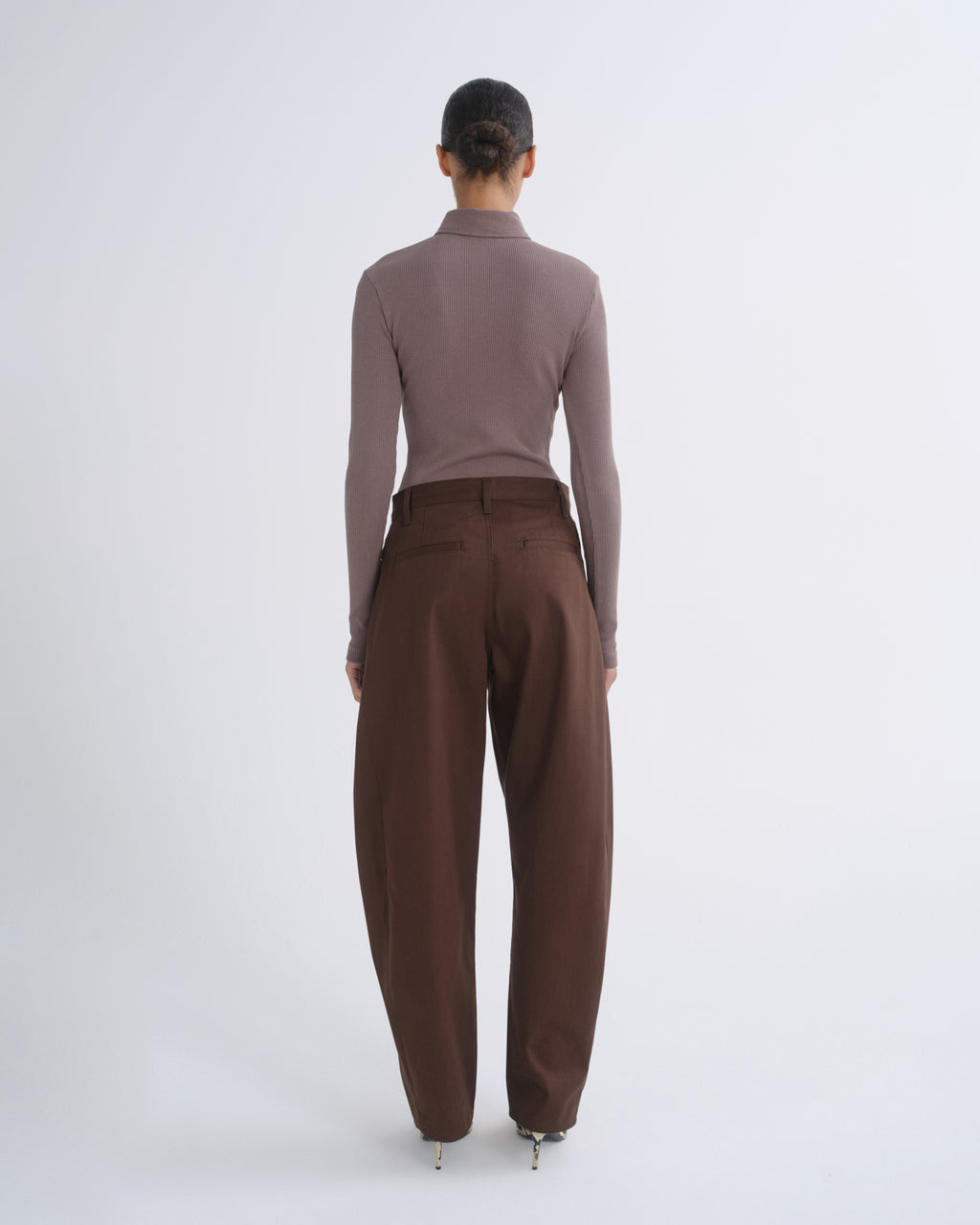 A model shown from the back wearing a taupe-brown mock neck long-sleeve top and dark brown wide-leg trousers, standing against a light gray studio background.