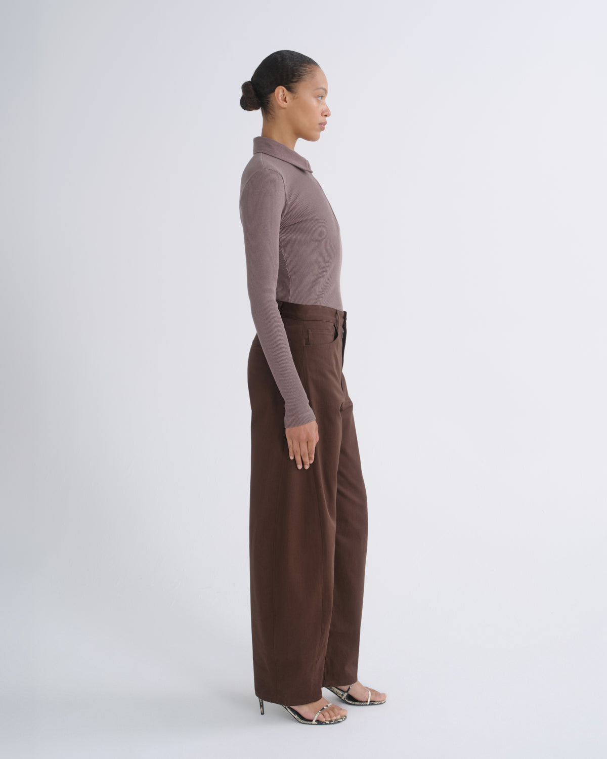 A woman stands in profile wearing a fitted long-sleeve polo-neck top in taupe and wide-leg brown trousers, photographed against a clean light gray studio background.