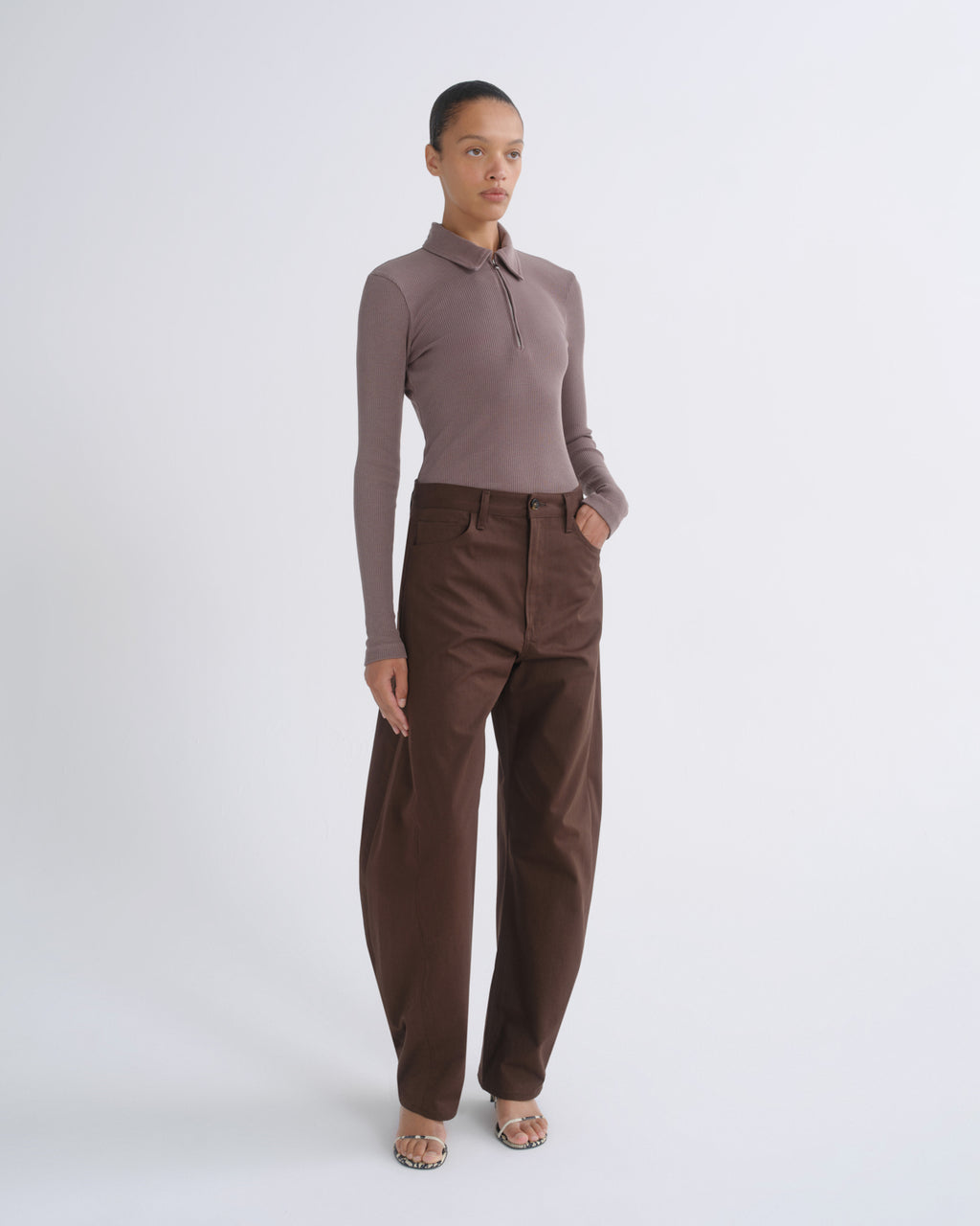 A model stands against a light gray studio background wearing a fitted long-sleeve polo top in taupe and high-waisted brown trousers.