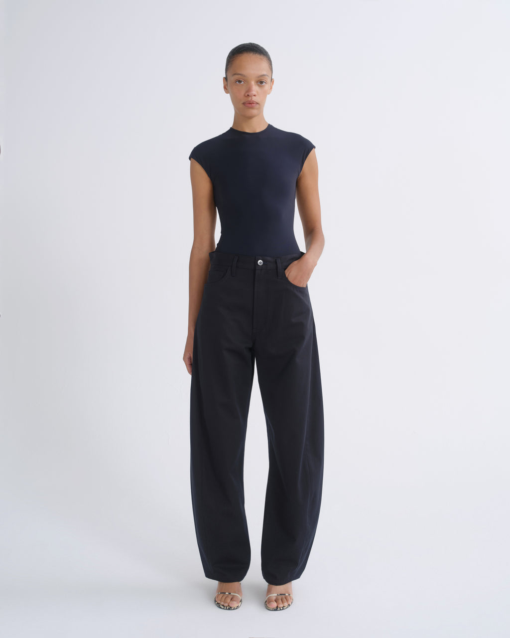 A model stands against a light gray studio background wearing a dark navy sleeveless mock-neck top and matching dark navy wide-leg pants.