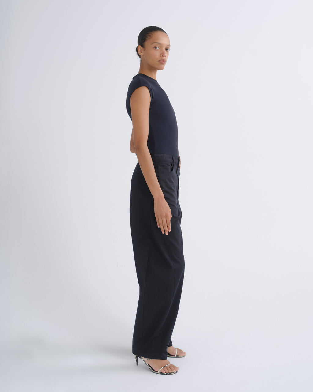 A model stands in profile wearing a sleeveless dark navy top and matching dark navy wide-leg trousers, paired with clear-strap high-heeled sandals, against a light gray studio background.