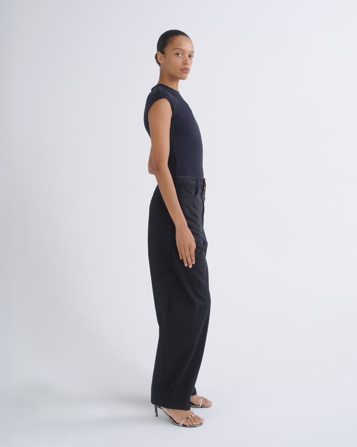 A model stands in profile wearing a sleeveless dark navy top and matching dark navy wide-leg trousers, paired with clear-strap high-heeled sandals, against a light gray studio background.