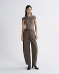 A model stands against a light gray studio background wearing a taupe-brown short-sleeve top with a gathered, pleated yoke and matching wide-leg trousers, paired with dark brown heeled boots.