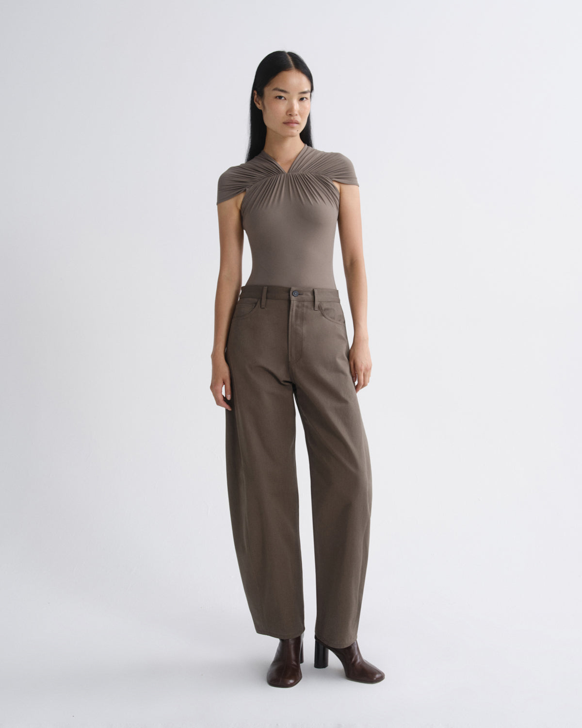 A model stands against a light gray studio background wearing a taupe-brown short-sleeve top with a gathered, pleated yoke and matching wide-leg trousers, paired with dark brown heeled boots.
