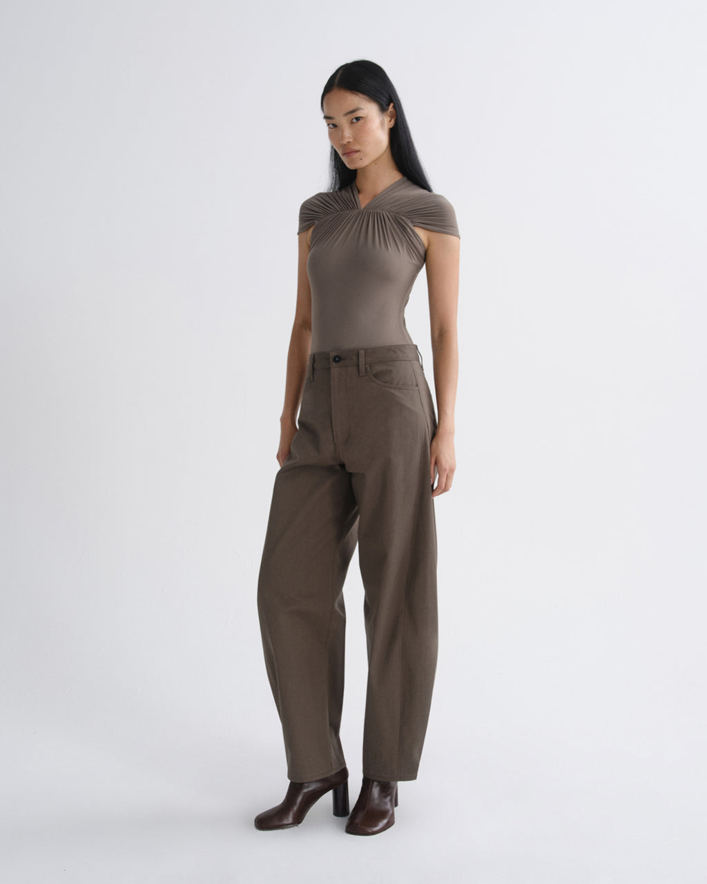 A woman modeling a taupe brown short-sleeve top with a gathered neckline and matching wide-leg trousers, paired with dark brown heeled boots, against a light gray studio background.