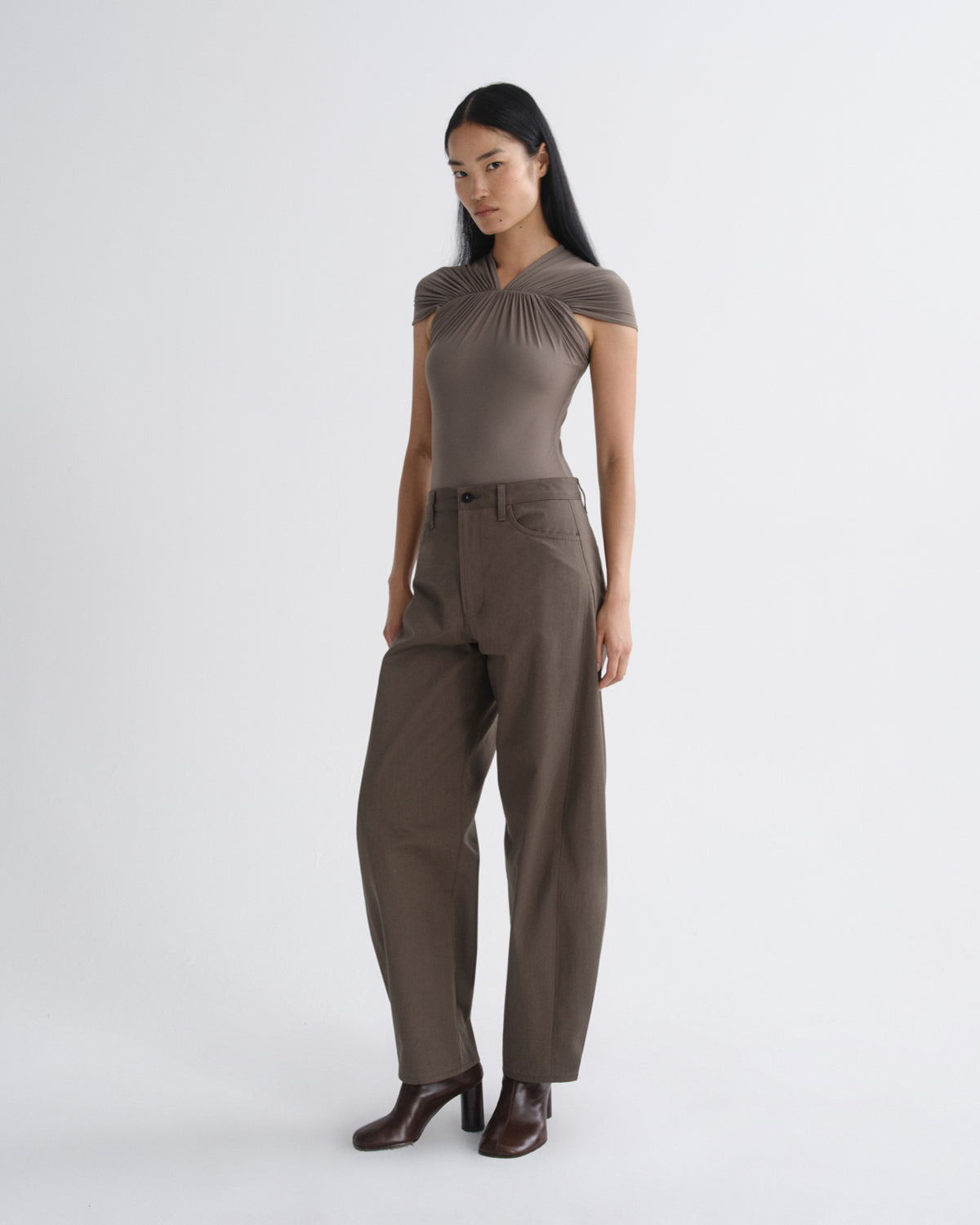 A woman modeling a taupe brown short-sleeve top with a gathered neckline and matching wide-leg trousers, paired with dark brown heeled boots, against a light gray studio background.
