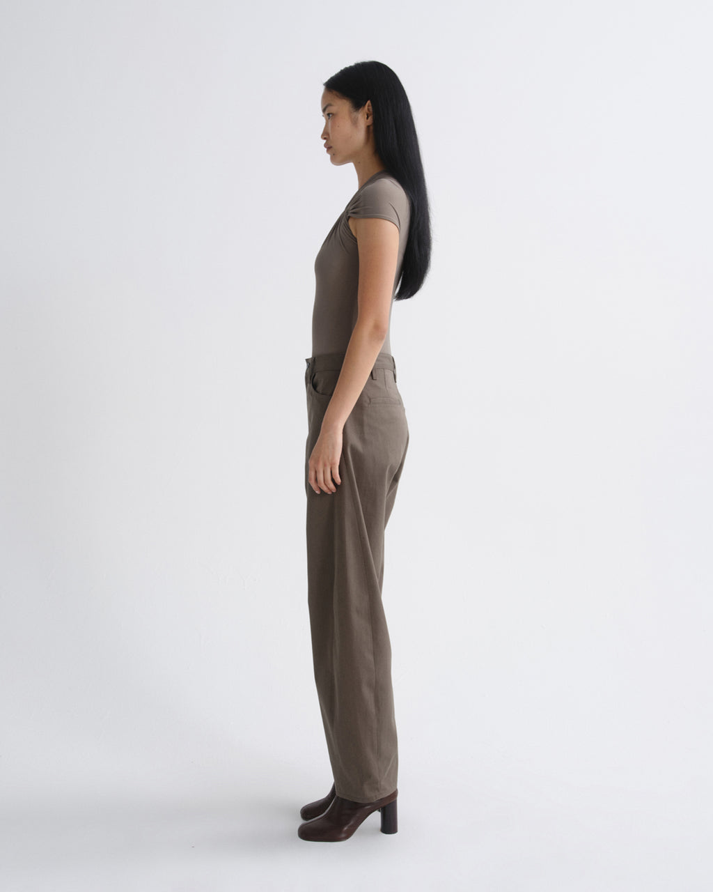 A woman in a side profile view wearing a taupe short-sleeve top and matching wide-leg trousers, paired with dark brown heeled shoes, against a plain light gray studio background.