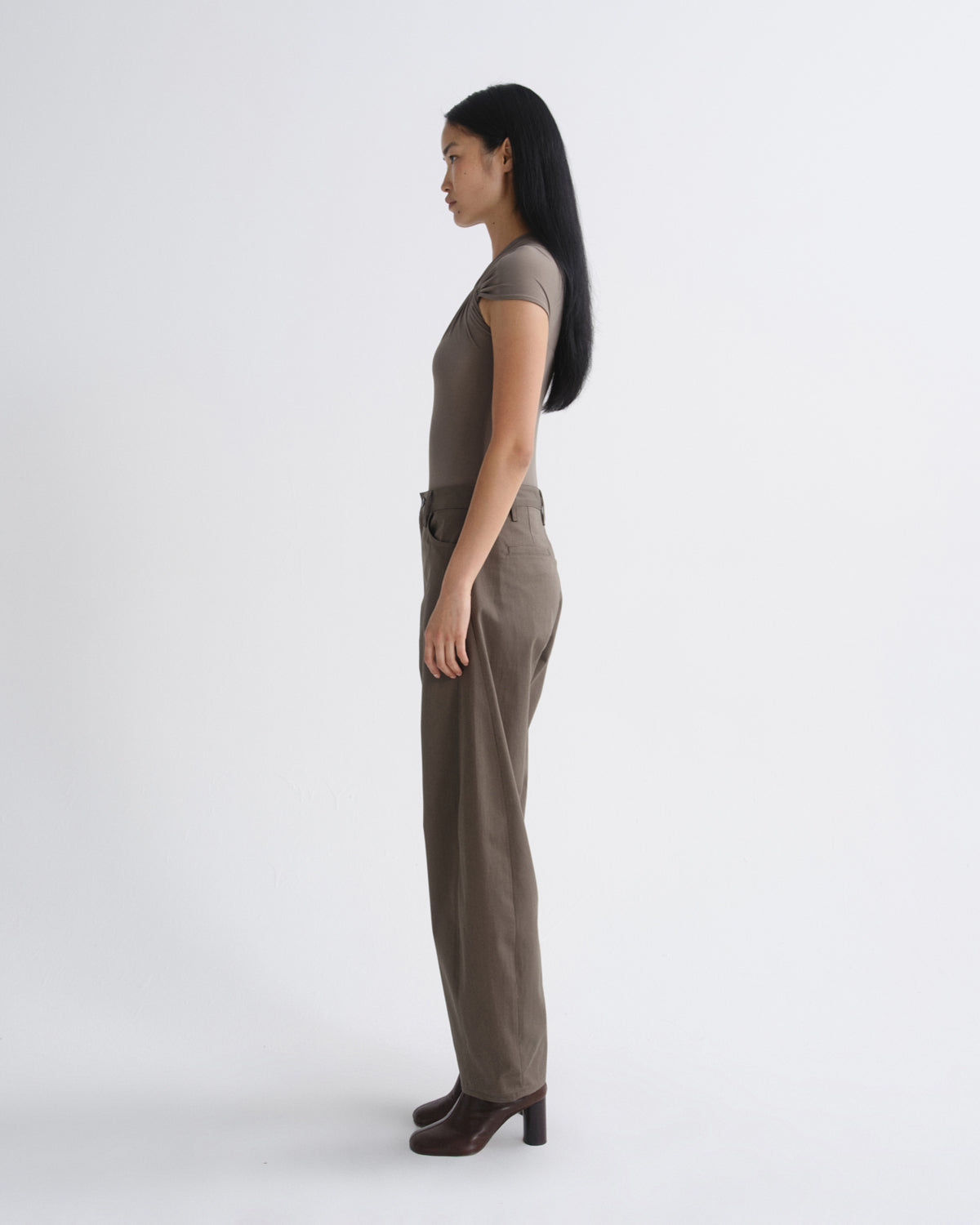 A woman in a side profile view wearing a taupe short-sleeve top and matching wide-leg trousers, paired with dark brown heeled shoes, against a plain light gray studio background.