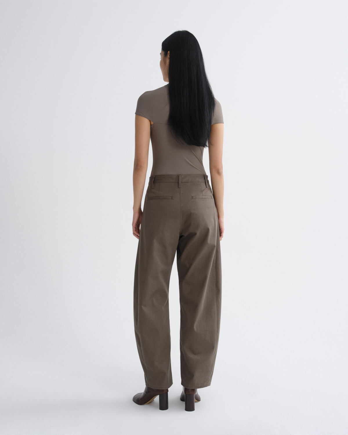 A woman with long black hair is shown from behind wearing a taupe short-sleeve top and matching wide-leg trousers, paired with dark brown block-heel shoes, against a plain light gray studio background.