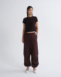 A model stands against a light gray studio background wearing a dark brown short-sleeve crop top, dark brown wide-leg cargo pants, and white pointed shoes.