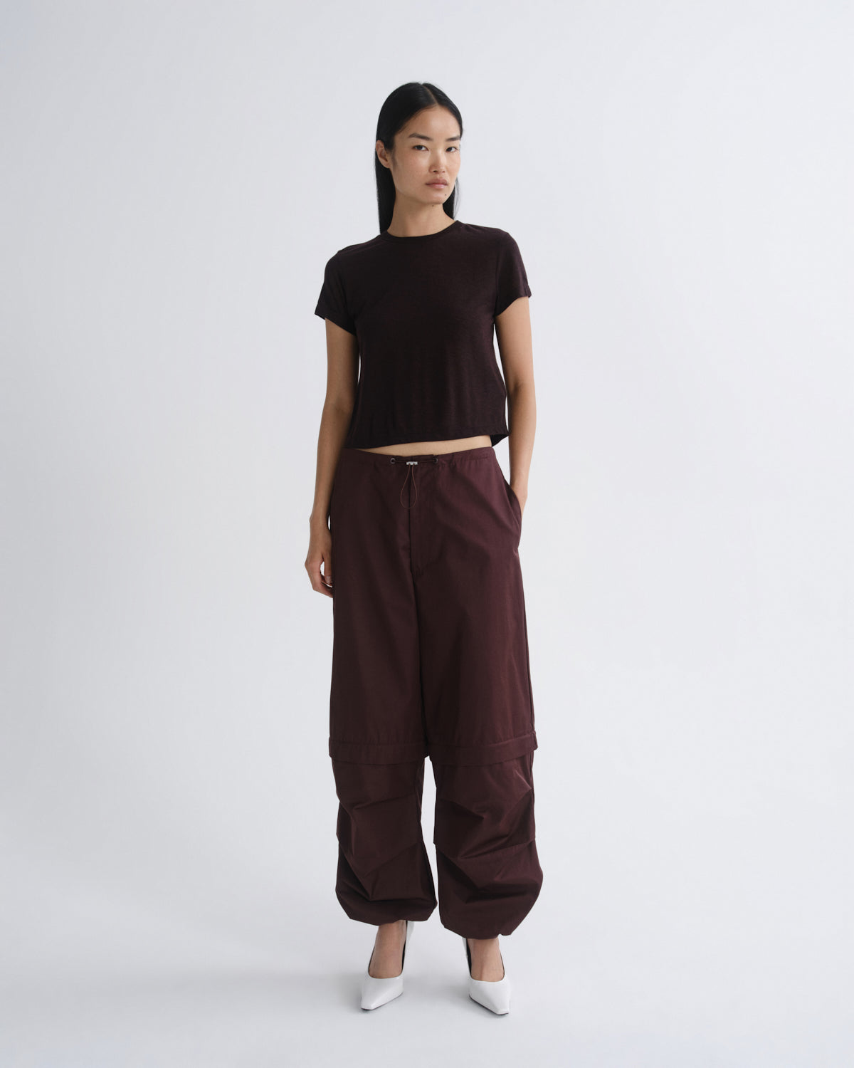 A model stands against a light gray studio background wearing a dark brown short-sleeve crop top, dark brown wide-leg cargo pants, and white pointed shoes.