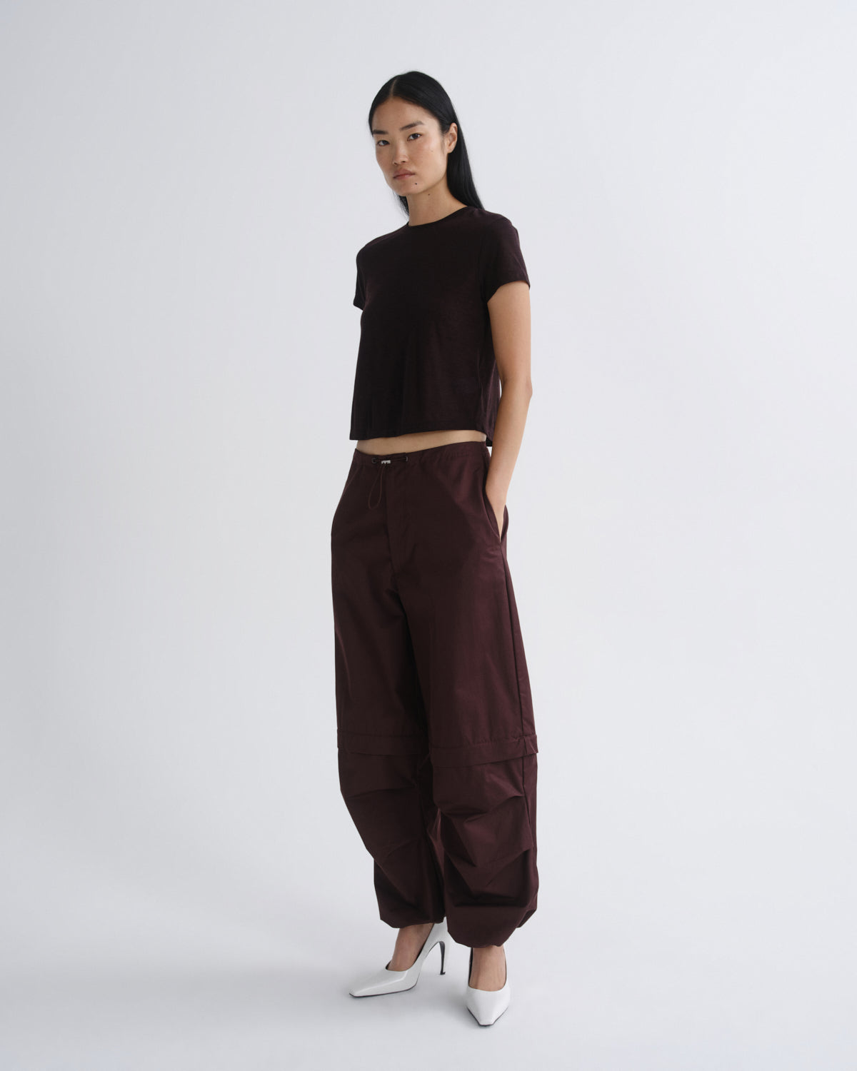 A model stands against a light gray studio background wearing a dark brown short-sleeve crop top, matching dark brown wide-leg cargo pants, and white pointed-toe heels.
