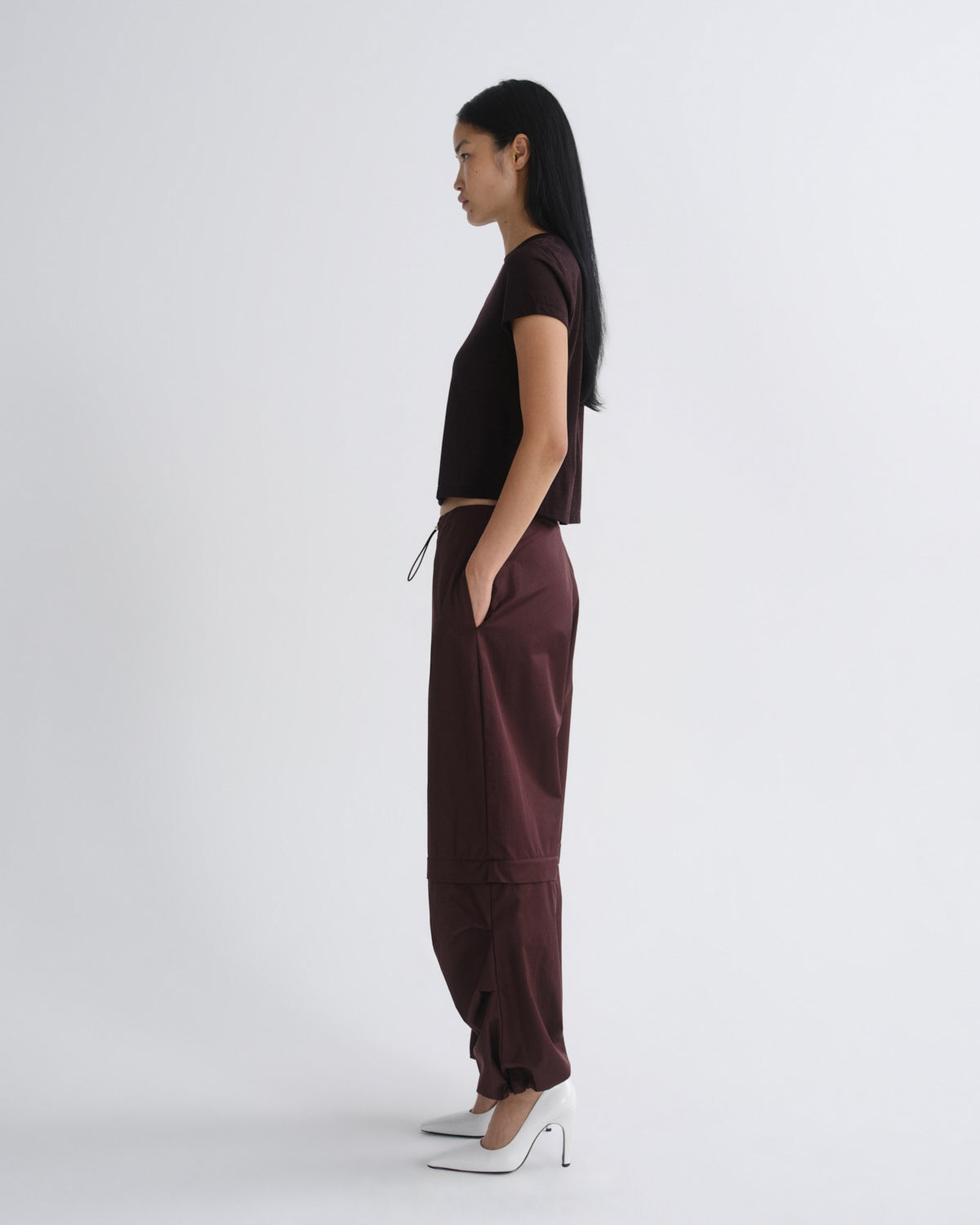 A woman stands in profile wearing a dark brown short-sleeve crop top and matching dark brown wide-leg pants with white pointed heels, photographed against a clean light gray studio background.