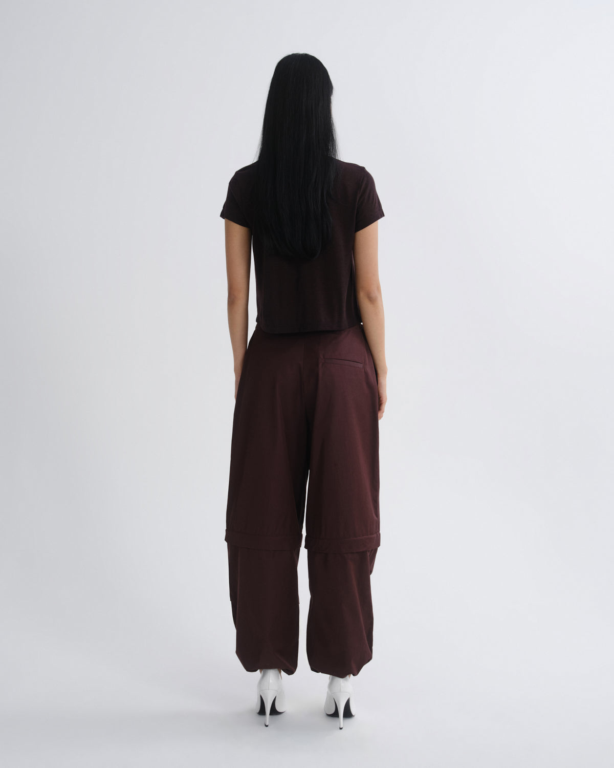 A model viewed from the back wearing a dark brown short-sleeve t-shirt and matching dark brown wide-leg cargo pants, paired with white high-heeled shoes, against a light gray studio background.