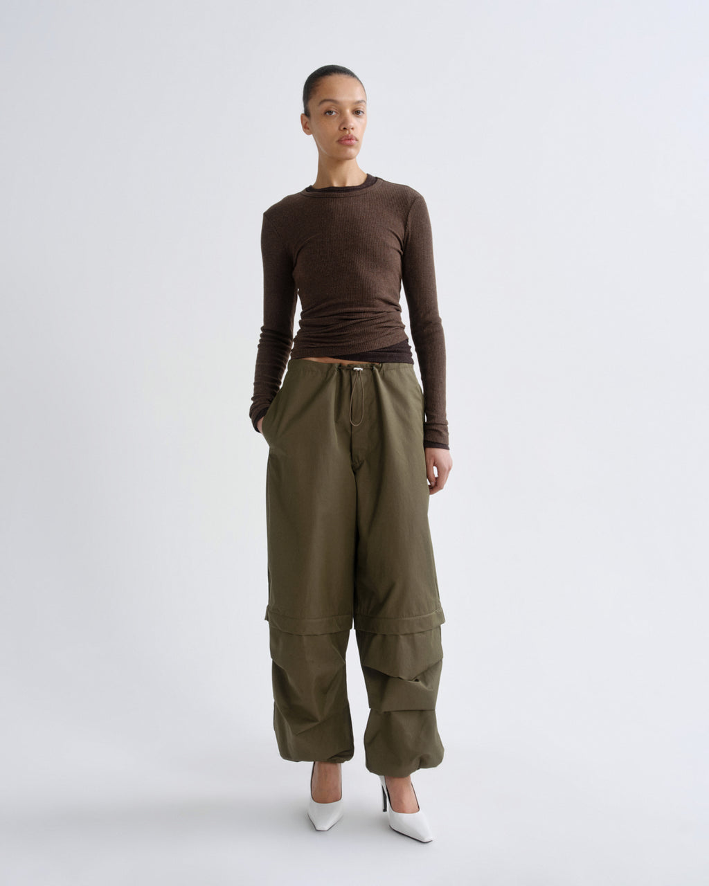 A model stands against a light gray studio background wearing a fitted brown long-sleeve top, olive green wide-leg cargo pants, and white pointed shoes.