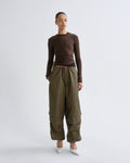A model stands against a light gray studio background wearing a fitted brown long-sleeve top, olive green wide-leg cargo pants, and white pointed shoes.