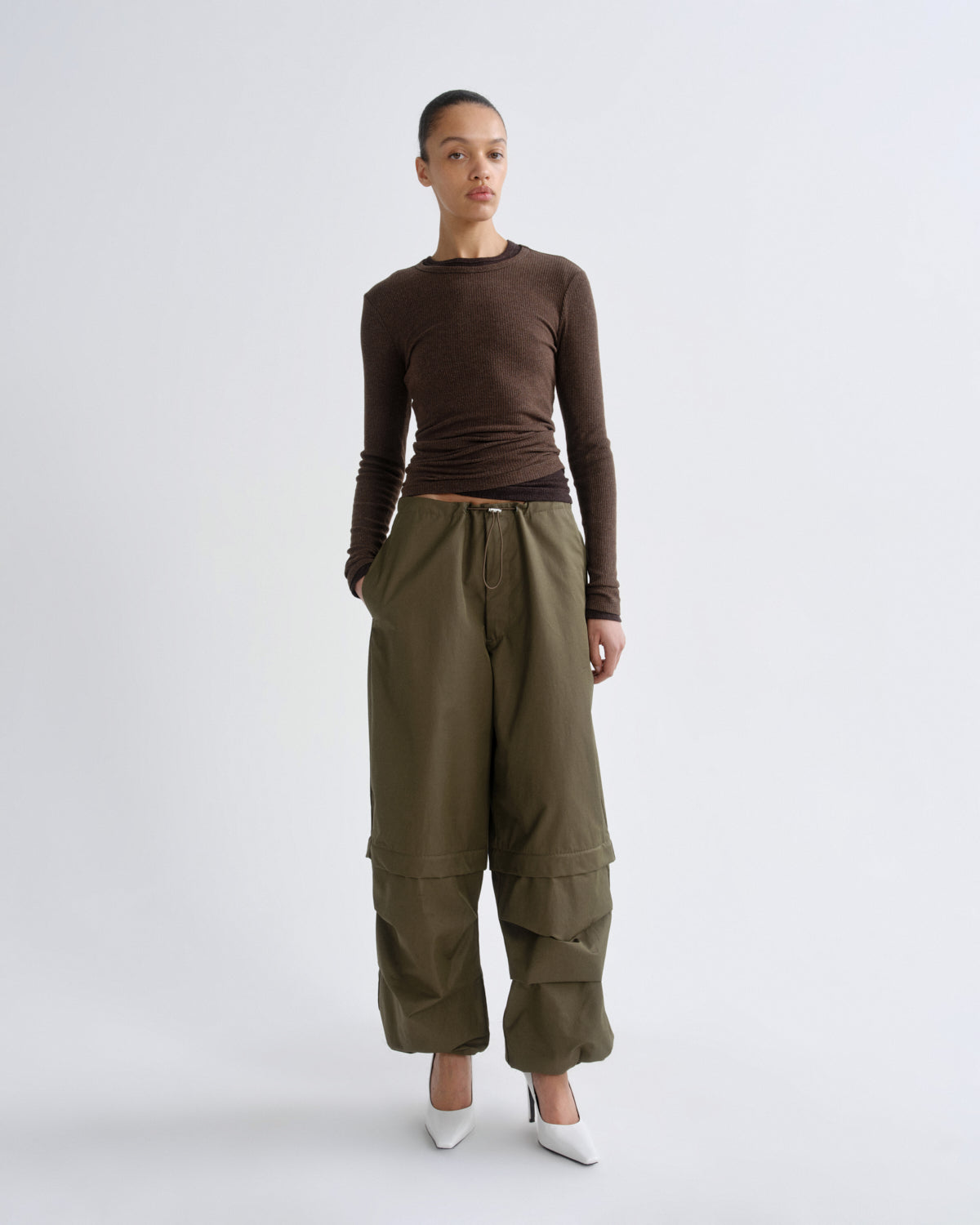 A model stands against a light gray studio background wearing a fitted brown long-sleeve top, olive green wide-leg cargo pants, and white pointed shoes.