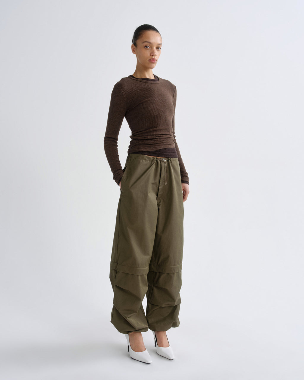 A model stands against a light gray studio background wearing a fitted brown long-sleeve top, loose olive green wide-leg pants, and white pointed shoes.