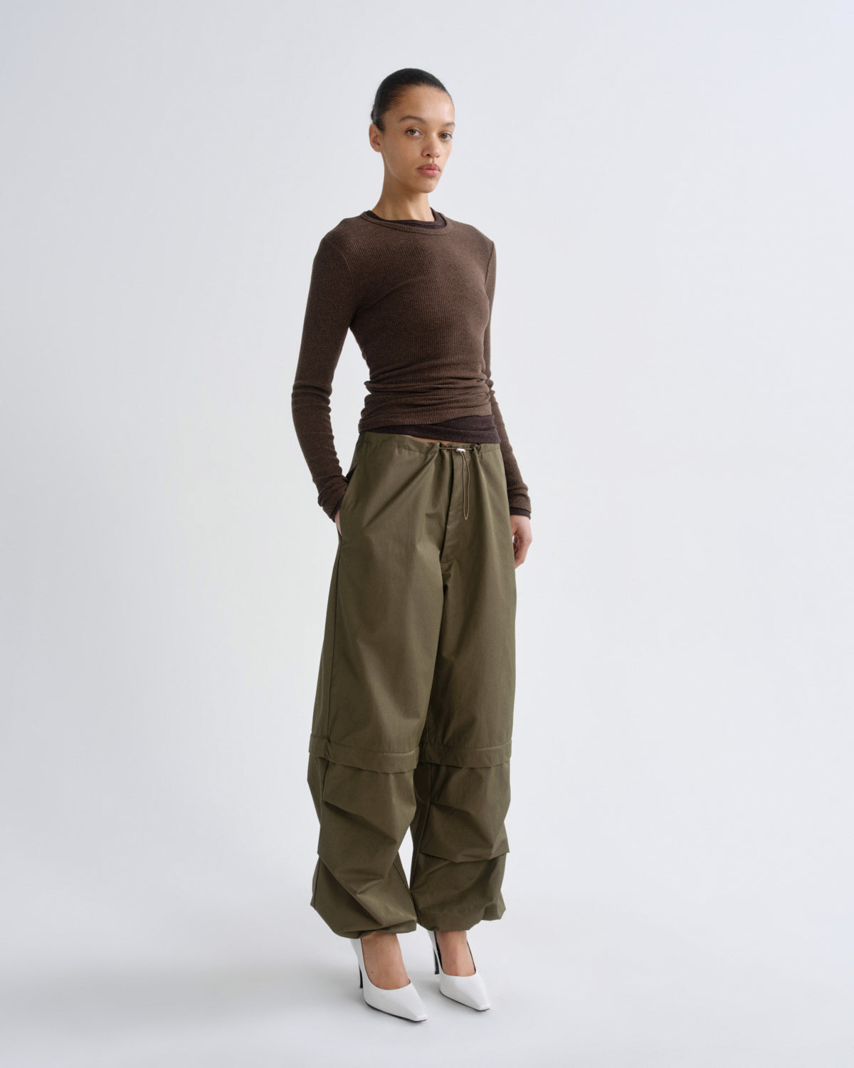 A model stands against a light gray studio background wearing a fitted brown long-sleeve top, loose olive green wide-leg pants, and white pointed shoes.