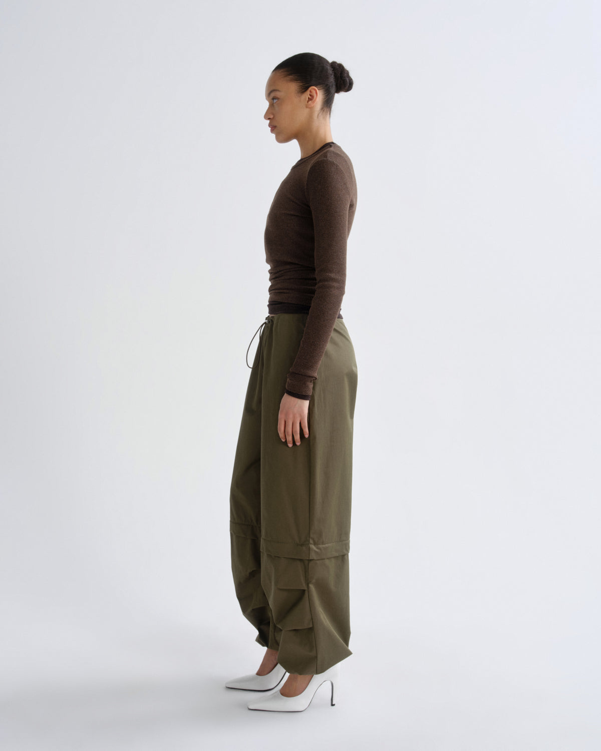 A woman stands in profile wearing a fitted brown long-sleeve top, olive green wide-leg cargo pants, and white pointed heels against a plain light gray studio background.