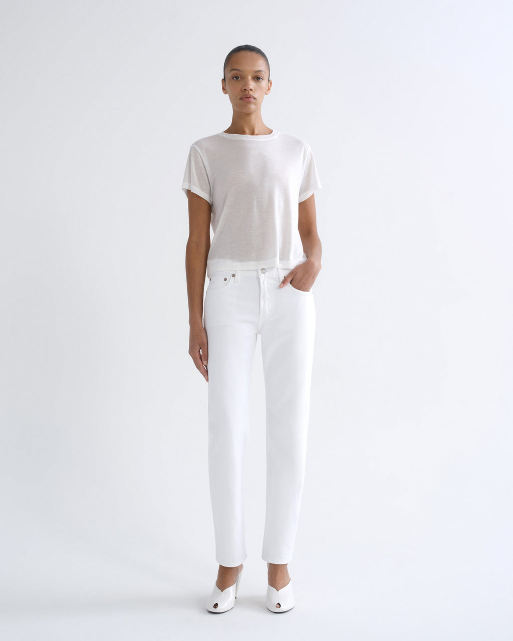 A model stands against a plain white studio background wearing a loose white short-sleeve top, white straight-leg jeans, and white pointed shoes.