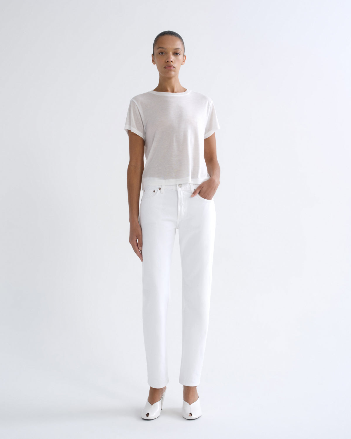 A model stands against a plain white studio background wearing a loose white short-sleeve top, white straight-leg jeans, and white pointed shoes.