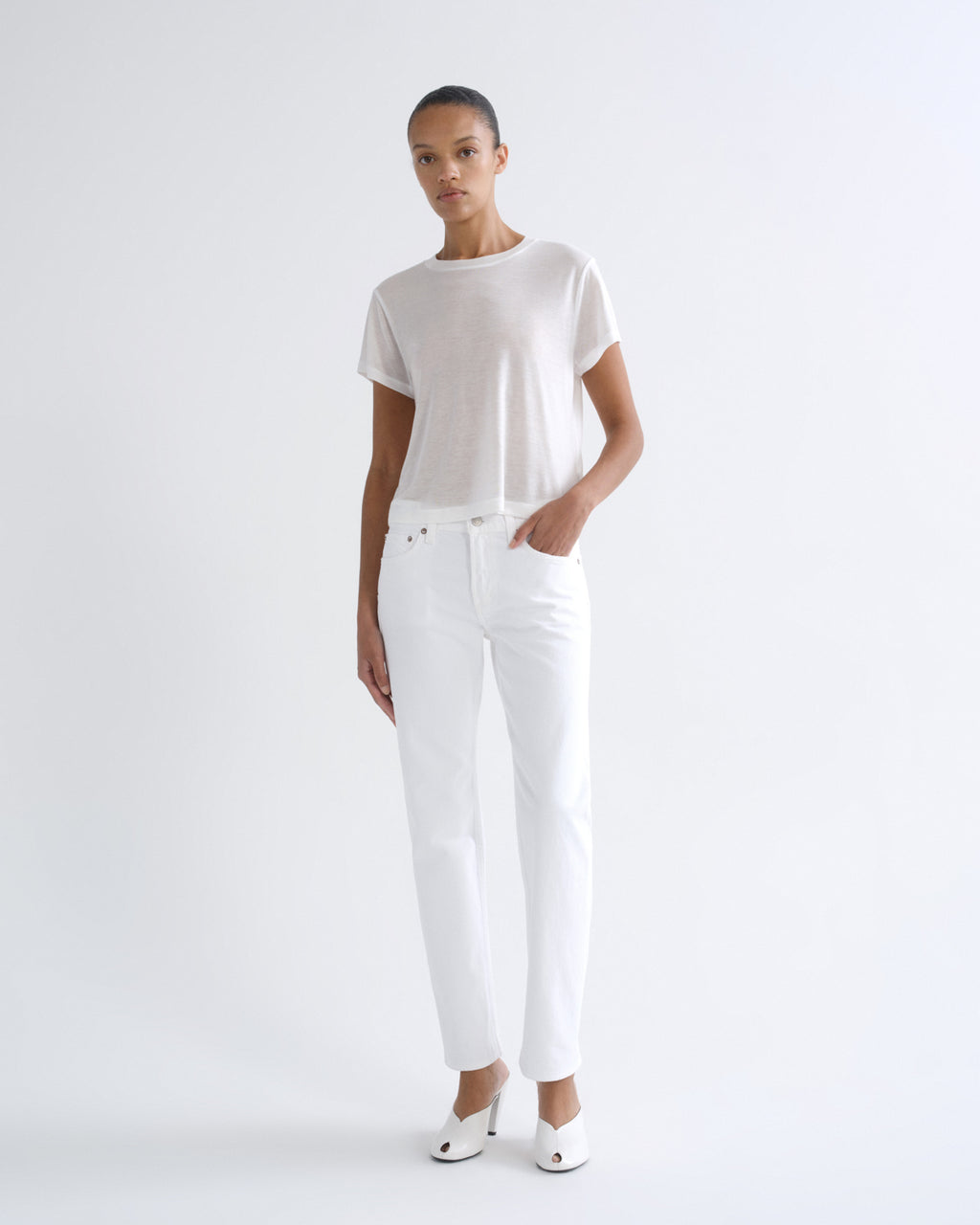 A model stands against a seamless white studio background wearing a white short-sleeve knit top, white straight-leg jeans, and white pointed-toe heels.