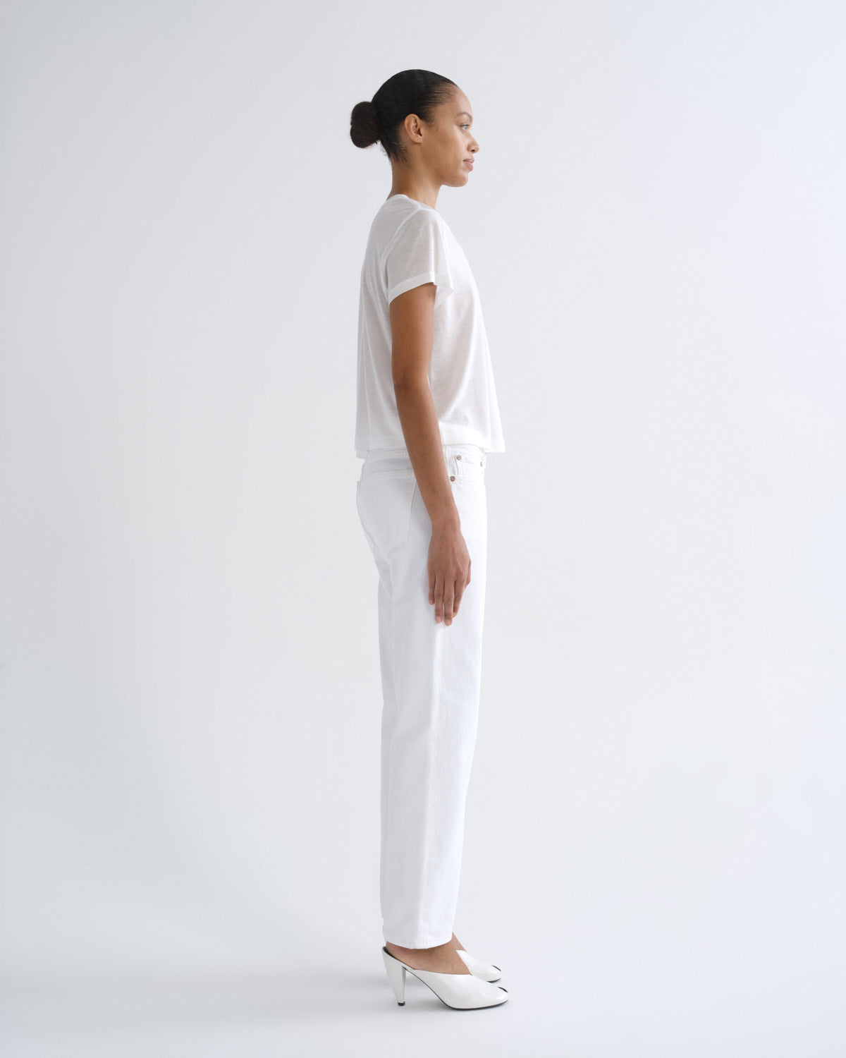 A woman in a side profile wearing a white short-sleeve top, white trousers, and white pointed-toe mule heels against a plain light gray studio background.