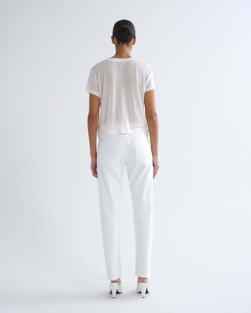 A model viewed from the back wearing a white short-sleeve crewneck t-shirt, white trousers, and white high heels against a plain white studio background.