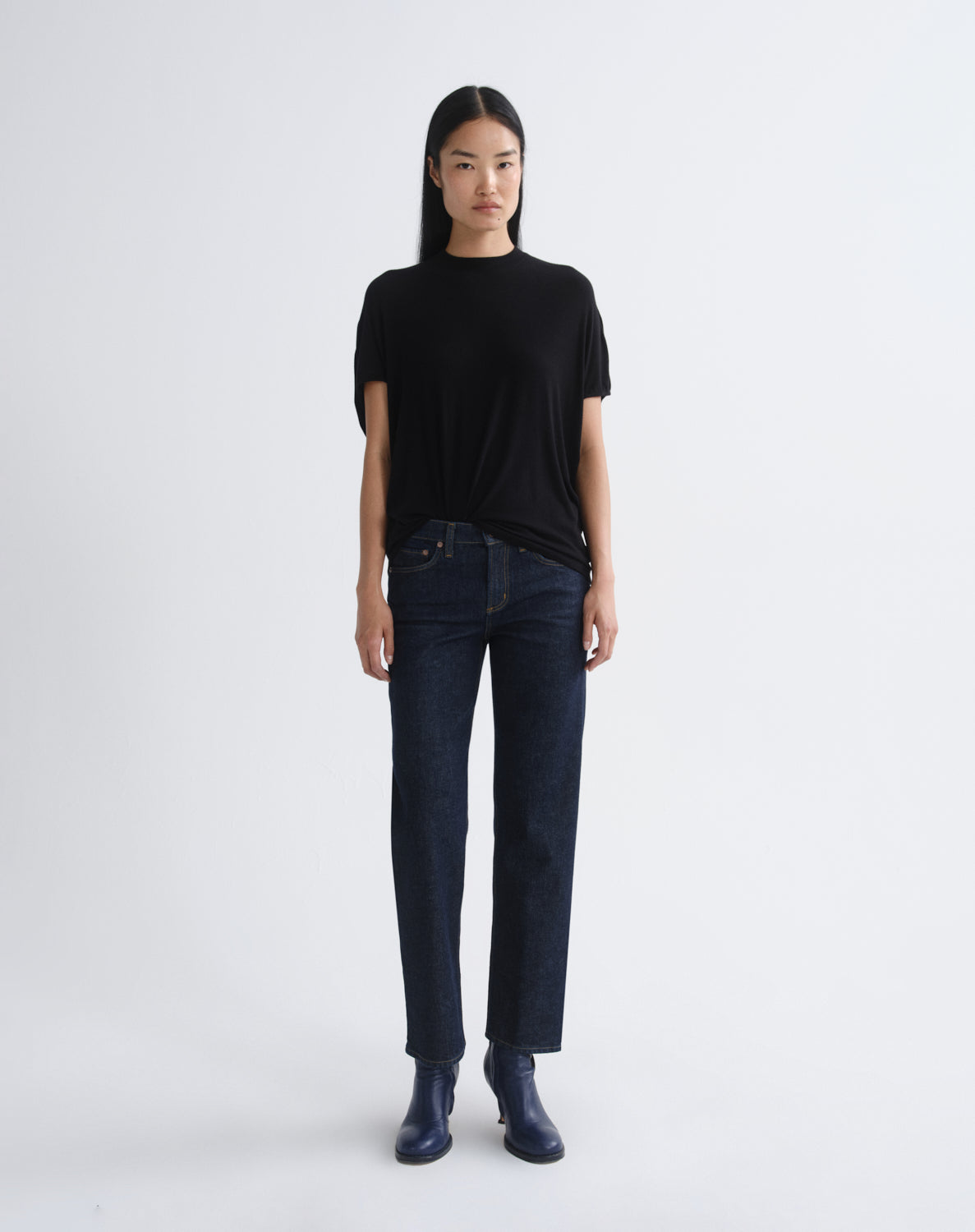 A model stands against a light gray studio background wearing a black short-sleeve t-shirt, dark blue straight-leg jeans, and dark blue boots.
