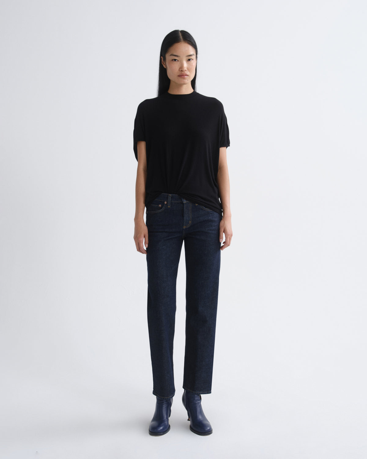 A model stands against a light gray studio background wearing a black short-sleeve t-shirt, dark blue straight-leg jeans, and dark blue heeled boots.
