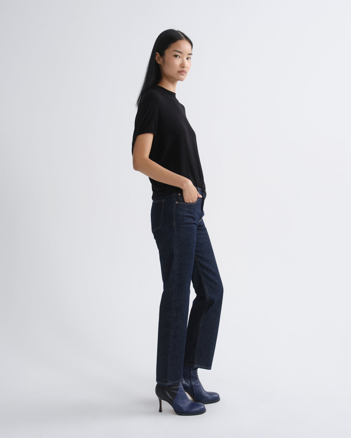 A woman stands in profile wearing a black short-sleeve top, dark blue straight-leg jeans, and dark blue heeled ankle boots against a light gray studio background.