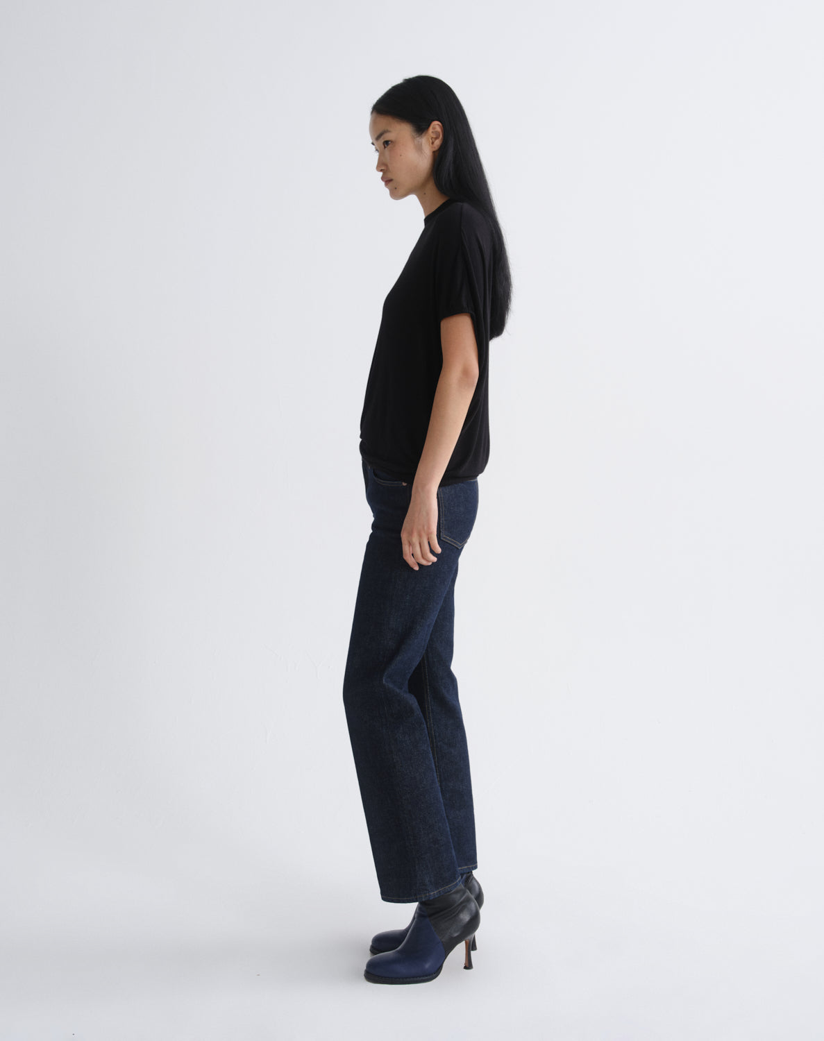 A model in side profile wearing a black short-sleeve top, dark blue wide-leg jeans, and dark blue heeled ankle boots, standing against a light gray studio background.