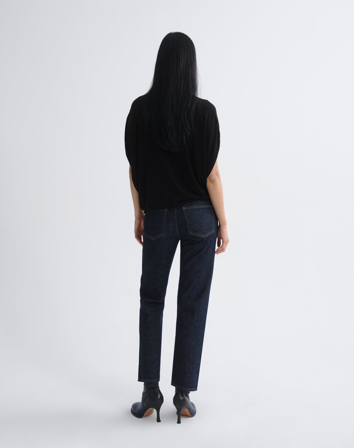 A woman with long black hair is shown from behind wearing a black short-sleeve top, dark blue straight-leg jeans, and black ankle boots, standing against a light gray studio background.
