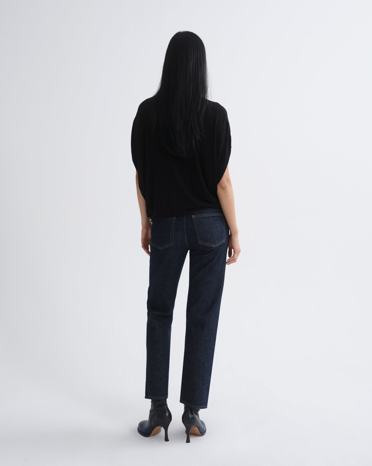 A woman is shown from the back wearing a black short-sleeve top, dark blue straight-leg jeans, and black high-heeled boots against a plain light gray studio background.